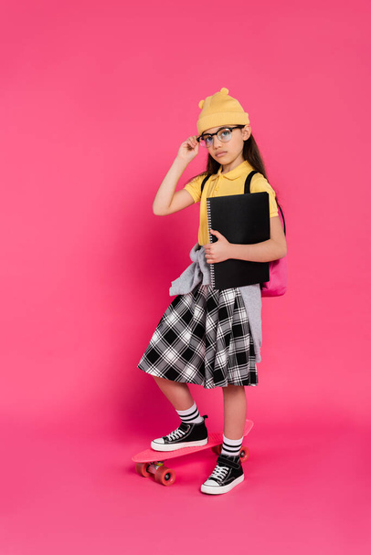 schoolgirl in beanie hat and glasses holding notebooks, standing near penny board on pink background - Photo, Image