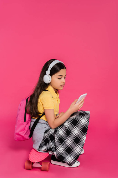 colegiala en auriculares inalámbricos sentado en el tablero de penique, fondo rosa, utilizando el teléfono inteligente - Foto, Imagen