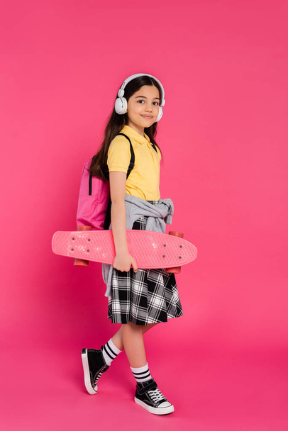 happy schoolgirl in wireless headphones standing with penny board, pink background, after classes - Photo, Image