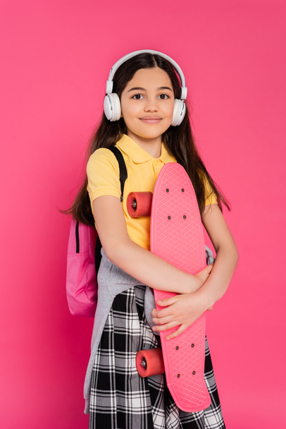 chica positiva en auriculares inalámbricos de pie con penny board, fondo rosa, después de clases - Foto, Imagen