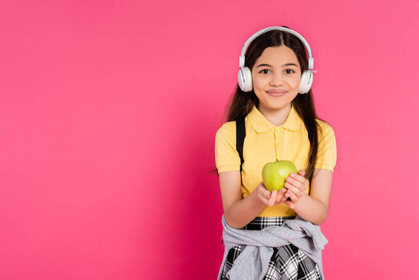 happy schoolgirl in wireless headphones holding green apple on pink background, kid with backpack - Photo, Image