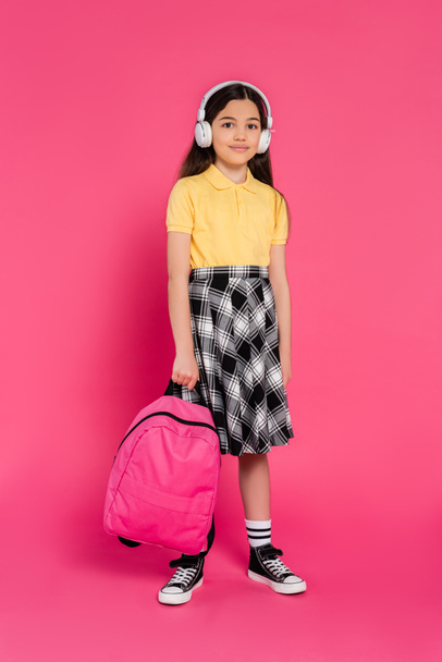 smiling schoolgirl in wireless headphones holding backpack on pink background, brunette student - Foto, Imagem