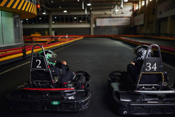 two diverse racers in helmets and sportswear driving go kart on indoor circuit, competitors - Photo, Image