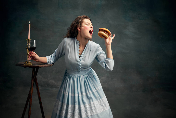 Comida rápida. Princesa. Retrato de mujer aristocrática vestida con vestido histórico azul comiendo hamburguesa grande. Arte gastronómico contemporáneo. Concepto de estilo de vida saludable, dieta, estilo, moda. - Foto, imagen