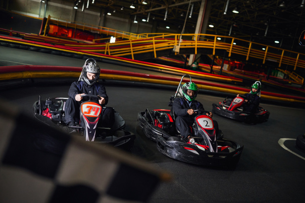 interracial men driving go kart near checkered black and white racing flag on blurred foreground - Photo, Image
