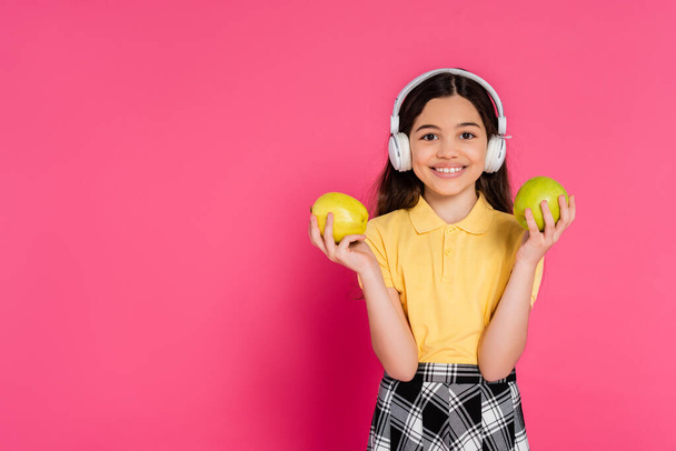colegiala sorprendida en auriculares inalámbricos sosteniendo manzana y mochila, fondo rosa, estudiante - Foto, Imagen