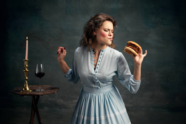 Een junkfood prinses. Portret van een aristocratische hongerige vrouw die blauwe historische jurk draagt en grote hamburger eet. Moderne voedselkunst. Concept van gezonde levensstijl, dieet, stijl, mode. - Foto, afbeelding