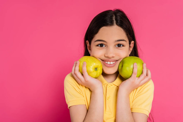 positive girl with brunette hair holding green apples on pink background, portrait, happiness - Photo, Image