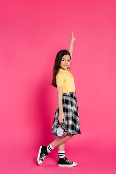 happy schoolgirl holding alarm clock on pink background, looking at camera, pointing up, full length - Photo, Image
