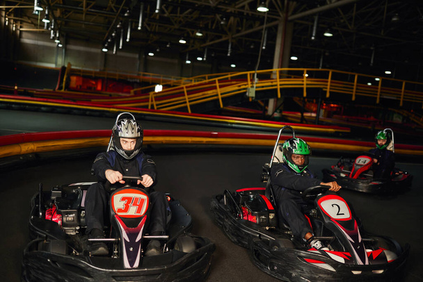 fokussierte multikulturelle Go-Kart-Rennfahrer auf Indoor-Rundkurs, Speed-Rennen und Motorsport - Foto, Bild