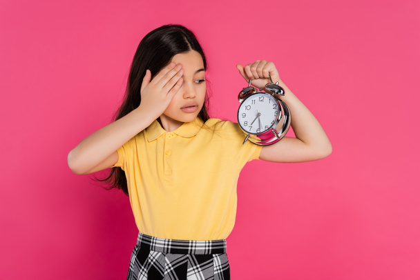 colegiala morena mirando reloj despertador vintage aislado en rosa, volver al concepto de la escuela, - Foto, Imagen