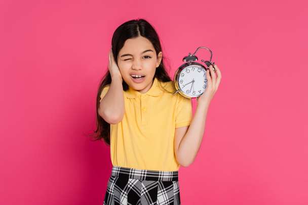 colegiala mirando a la cámara, guiñando el ojo, sosteniendo reloj despertador vintage aislado en rosa, de vuelta a la escuela - Foto, Imagen