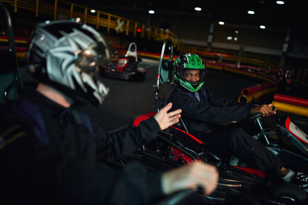 Africano americano go kart driver no capacete olhando para amigo e dirigindo no circuito, competição - Foto, Imagem
