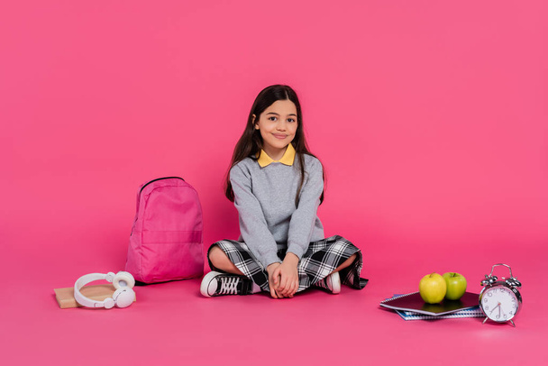 šťastná školačka sedící u batohu, zápisníky, sluchátka, zelená jablka, retro budík - Fotografie, Obrázek