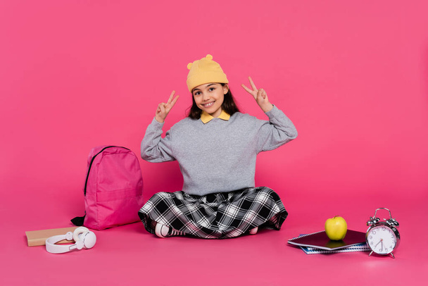 colegiala feliz en gorro sombrero mostrando v signo, cuadernos, auriculares, manzana, mochila, despertador - Foto, Imagen