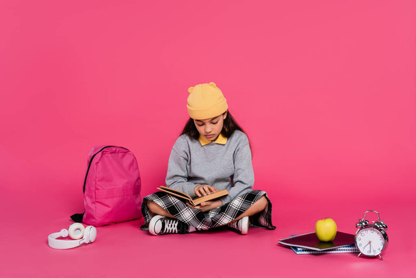 μαθήτρια σε Beanie καπέλο βιβλίο ανάγνωσης, κάθεται κοντά σε ακουστικά, μήλο, σακίδιο, ξυπνητήρι - Φωτογραφία, εικόνα