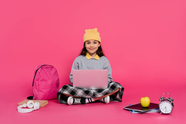 χαρούμενη μαθήτρια με καπέλο beanie που χρησιμοποιεί φορητό υπολογιστή, κάθεται κοντά σε ακουστικά, μήλο, σακίδιο, ξυπνητήρι - Φωτογραφία, εικόνα
