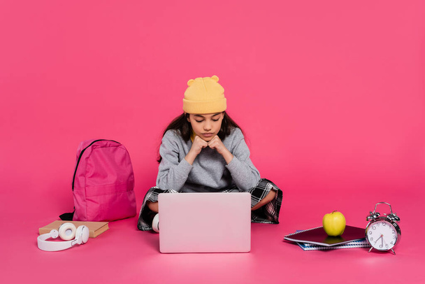 μαθητριούλα σε καπέλο beanie χρησιμοποιώντας φορητό υπολογιστή, κάθεται κοντά σε ακουστικά, πράσινο μήλο, σακίδιο, ξυπνητήρι - Φωτογραφία, εικόνα