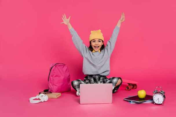 cheerful girl in beanie hat using laptop, sitting near headphones, apple, backpack, alarm clock - Photo, Image