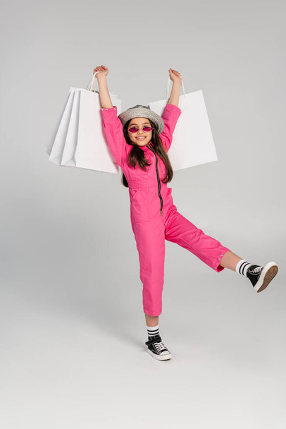 cheerful girl in stylish pink outfit and panama hat holding shopping bags on grey background - Foto, afbeelding