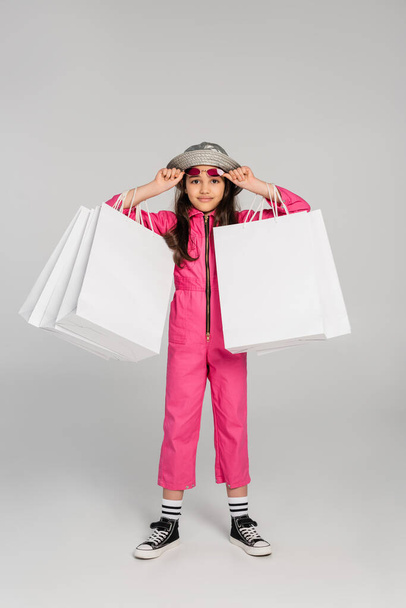 chica en traje elegante y sombrero panama sosteniendo bolsas de compras en gris, ajustando gafas de sol de color rosa - Foto, imagen