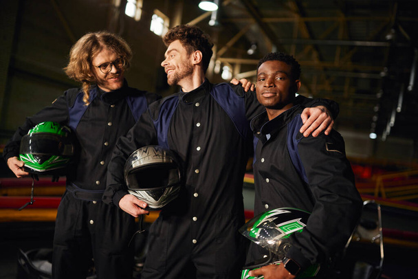 groep multiculturele en happy go kart chauffeurs in beschermende pakken knuffelen en houden helmen - Foto, afbeelding