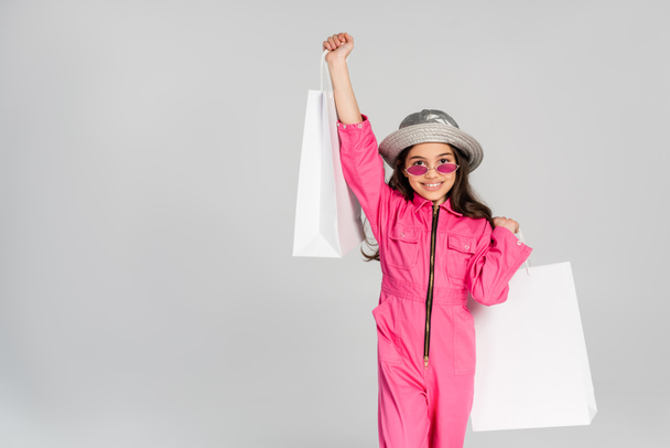 pozitivní dívka v módní oblečení, růžové sluneční brýle a panama klobouk pózovat s nákupními taškami na šedé - Fotografie, Obrázek