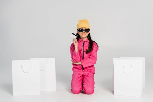 счастливая девушка в шапочке и солнцезащитных очках, держащая кредитку на серой, детские покупки, модный наряд - Фото, изображение