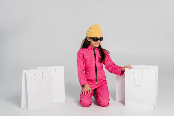 chica de moda en gorro sombrero y gafas de sol sentado cerca de bolsas de compras en gris, niño feliz, de moda - Foto, Imagen