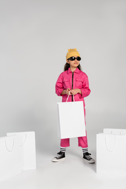 chica preadolescente con estilo en gorro sombrero y gafas de sol de pie y la celebración de bolsas de compras en gris - Foto, Imagen