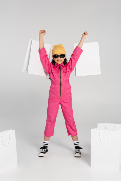 excited preteen girl in beanie hat and sunglasses holding shopping bags on grey background, style - Fotó, kép
