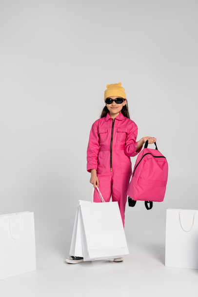 glückliches Schulmädchen mit Mütze und Sonnenbrille mit Einkaufstaschen und Rucksack auf grau, Stil - Foto, Bild