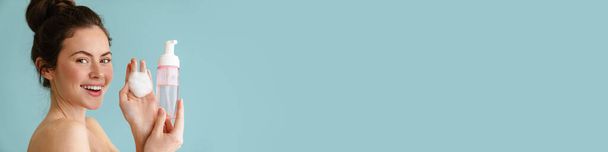 Pół naga brunetka uśmiecha się pokazując piankę czyszczącą odizolowaną na niebieskim tle - Zdjęcie, obraz