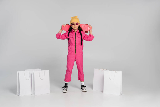 gelukkig meisje in beanie en stijlvolle zonnebril holding penny board, winkelen tassen op grijze achtergrond - Foto, afbeelding