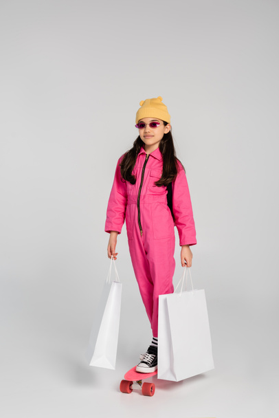 Stylisches Mädchen in Mütze und rosa Sonnenbrille reitet Penny Board und hält Einkaufstüten auf grau - Foto, Bild