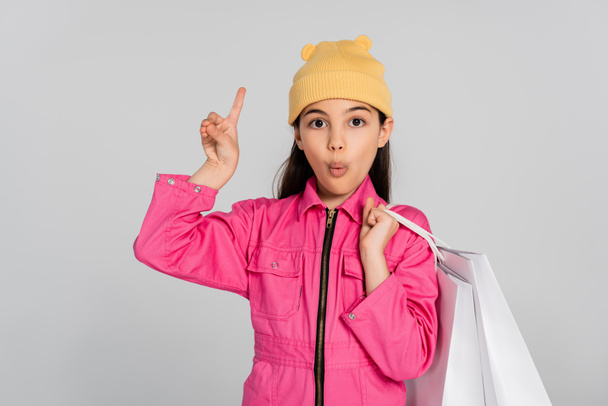 chica sorprendida en gorro sombrero y traje rosa sosteniendo bolsas de compras y apuntando hacia arriba, fondo gris - Foto, Imagen