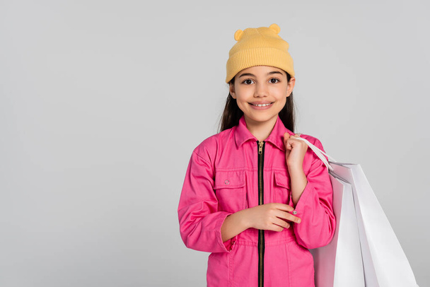 fröhliches Mädchen mit Mütze und rosa Outfit, Einkaufstaschen in der Hand und nach oben zeigend, grauer Hintergrund - Foto, Bild