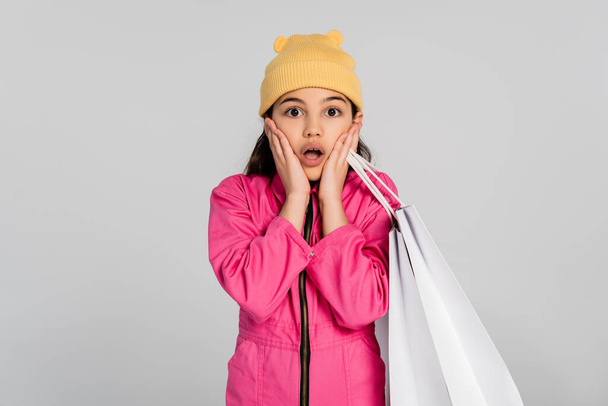 chica sorprendida en gorro sombrero y traje rosa sosteniendo bolsas de compras y mirando a la cámara, fondo gris - Foto, Imagen