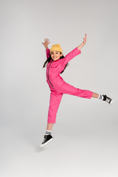 happy girl in beanie hat and stylish pink outfit jumping with outstretched hands on grey, have fun - Photo, Image