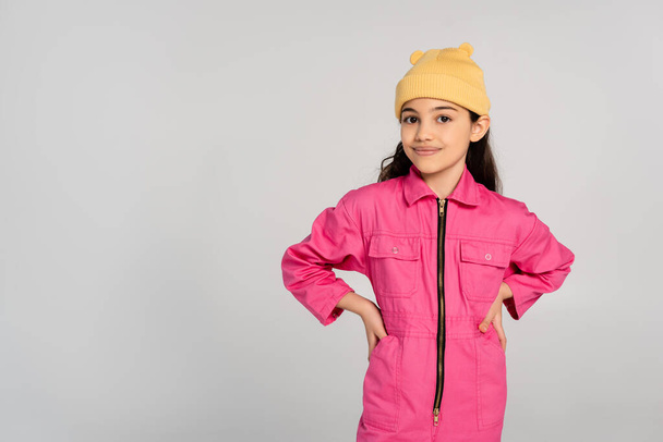 niño feliz en gorro amarillo sombrero y traje rosa de pie con las manos en las caderas aisladas en gris, moda - Foto, Imagen