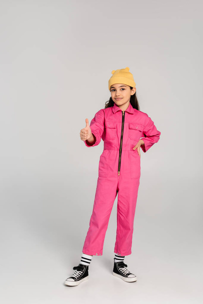 黄色いビーニー帽子とピンクの服の幸せな子供は,サイン,灰色の背景のようなヒップに手で立っています - 写真・画像