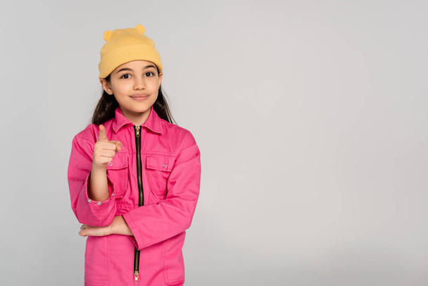 niño feliz en sombrero de gorro amarillo y traje rosa apuntando a la cámara en el fondo gris, niño con estilo - Foto, Imagen