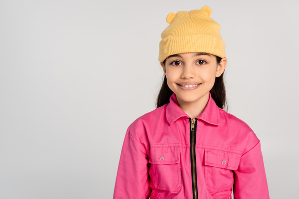 šťastná dívka ve žluté čepici a růžové oblečení při pohledu na kameru na šedém pozadí, stylové dítě - Fotografie, Obrázek