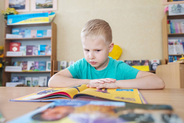 Een jongen in de bibliotheek. Zeven jaar Oekraïense man zit aan een tafel in de bibliotheek en boek te lezen. Concept van normaal leren, dorst naar kennis, verlangen om nieuwe dingen te leren. Wazige achtergrond en letters - Foto, afbeelding