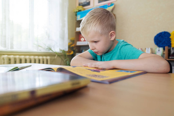 Хлопчик у бібліотеці. Сім років український хлопець сидить за столом у бібліотеці та читає книгу. Поняття нормального навчання, жага до знань, бажання вчитися нових речей. Розмитий фон і літери - Фото, зображення