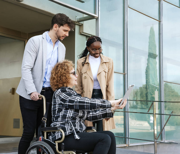 Adult businesswoman in wheelchair using a tablet with young businessmen outside an office buildingAdult businesswoman in wheelchair using a tablet with young businessmen outside an office building - Foto, Imagem