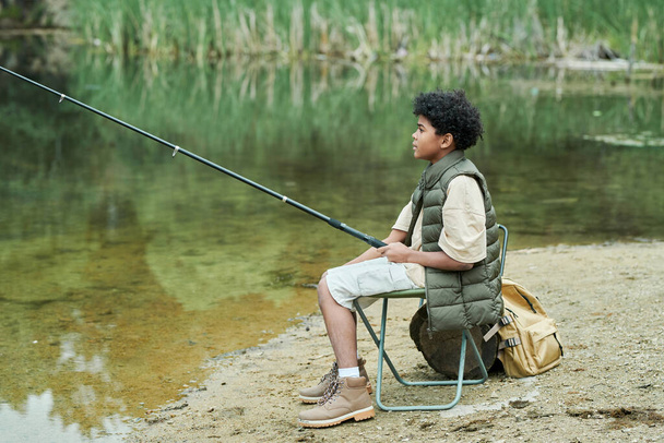 Παιδί που κάθεται σε καρέκλα με καλάμι ψαρέματος και ψαρεύει κοντά στη λίμνη - Φωτογραφία, εικόνα