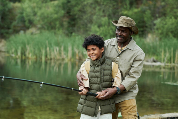 Happy African American dad catching fish together with his son on the lake outdoors - Photo, Image