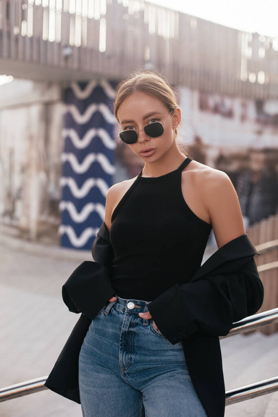 Model with trending clothes. Urban scene. Youth modeling. Urban trending fashion scene. Street fashion. Young gorgeous woman in black jacket, jeans and sunglasses  - Photo, Image