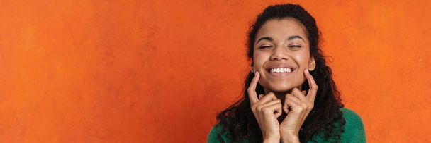 Happy smiling african casual brunette woman standing over orange wall background, grimacing, touching face - Foto, immagini